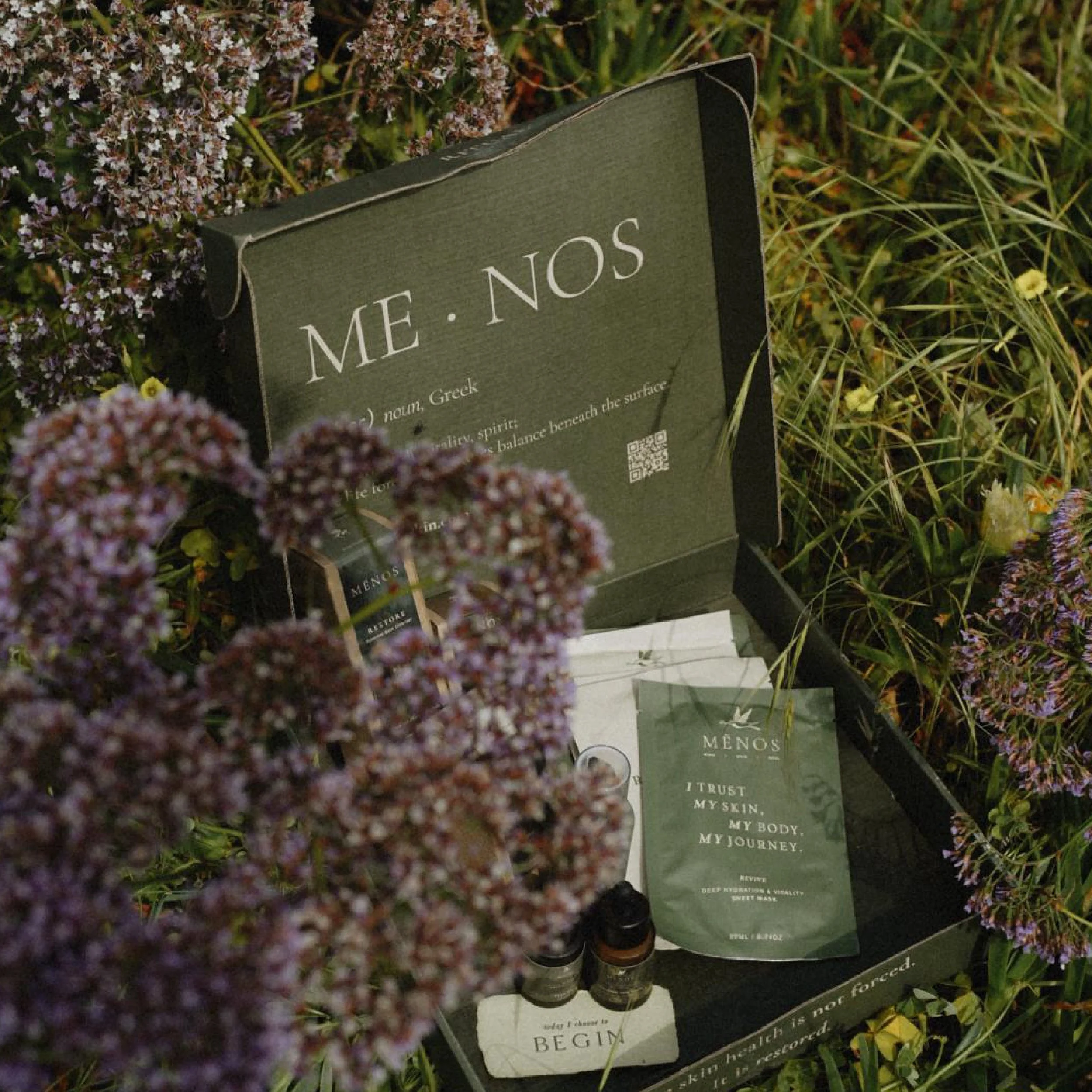 Open MĒNOS skincare gift box with serum and sachets, nestled among wild lilac flowers