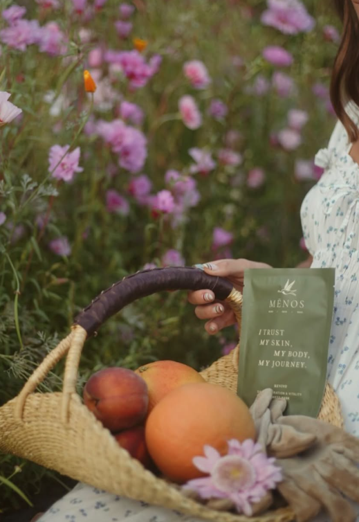 Woven basket with peaches and a MĒNOS sheet mask among pink cosmos flowers