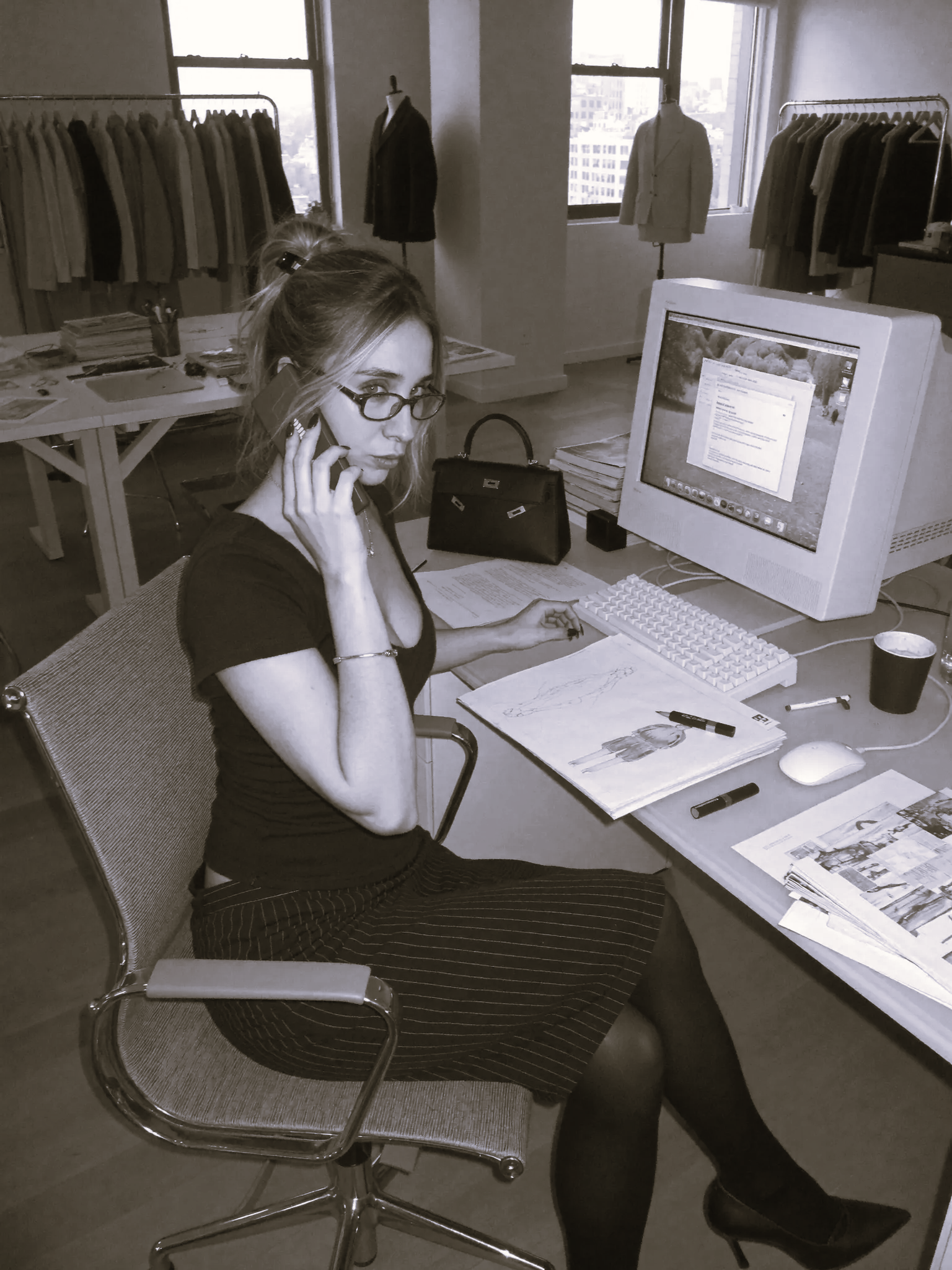 A woman with glasses talking on a landline phone at a cluttered desk with papers, magazines, and a computer monitor in an office with clothing racks and mannequins in the background.