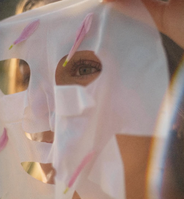 A person peering through a cutout of a sheet mask with pink and yellow design elements.