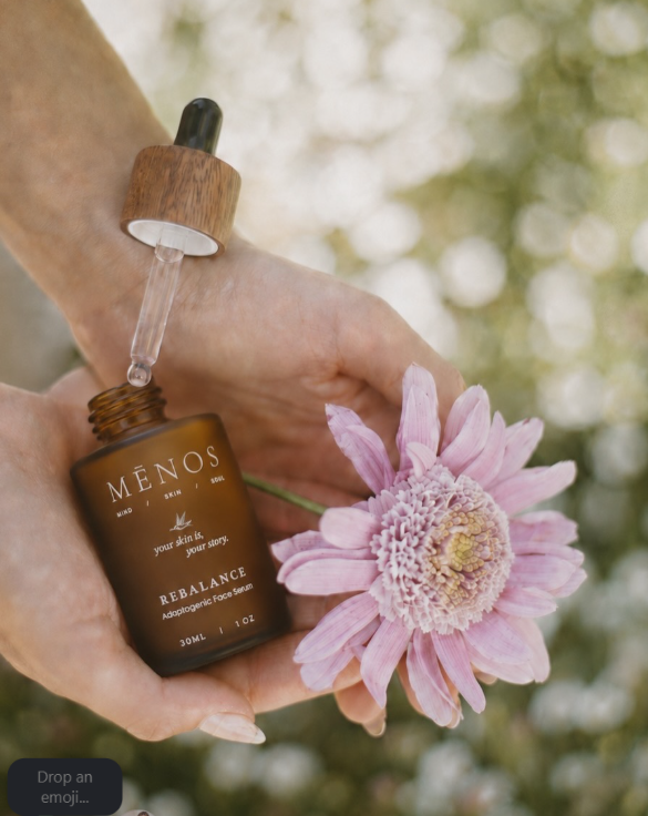 Hand holding the MĒNOS Rebalance serum bottle with its wooden dropper beside a pink flower