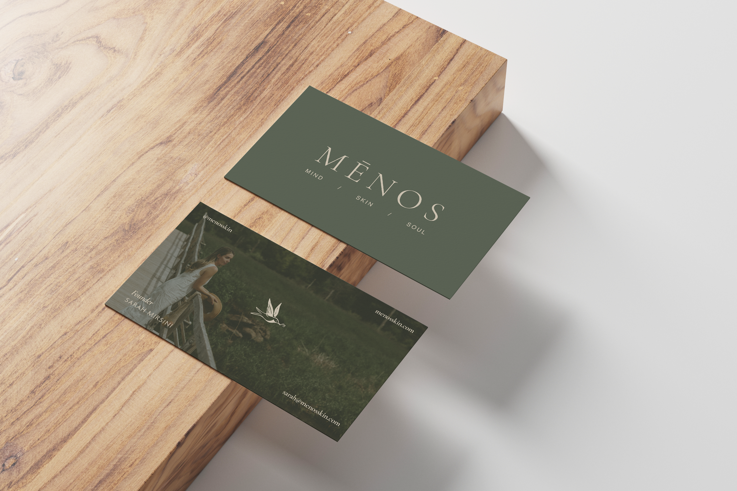 Sage green MĒNOS business cards, front and back, on a light oak block