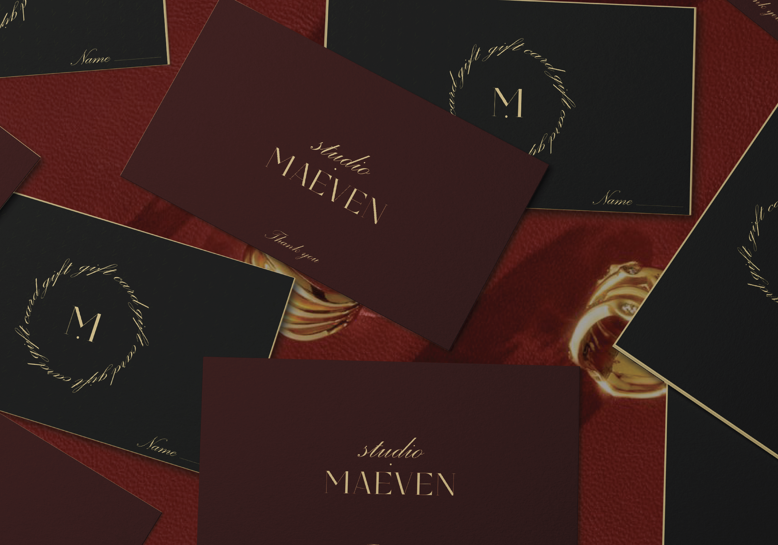 Multiple gift cards with maroon and black backgrounds, gold text, and the words 'studio MAEVEN' and a space for a name, arranged on a red textured surface.