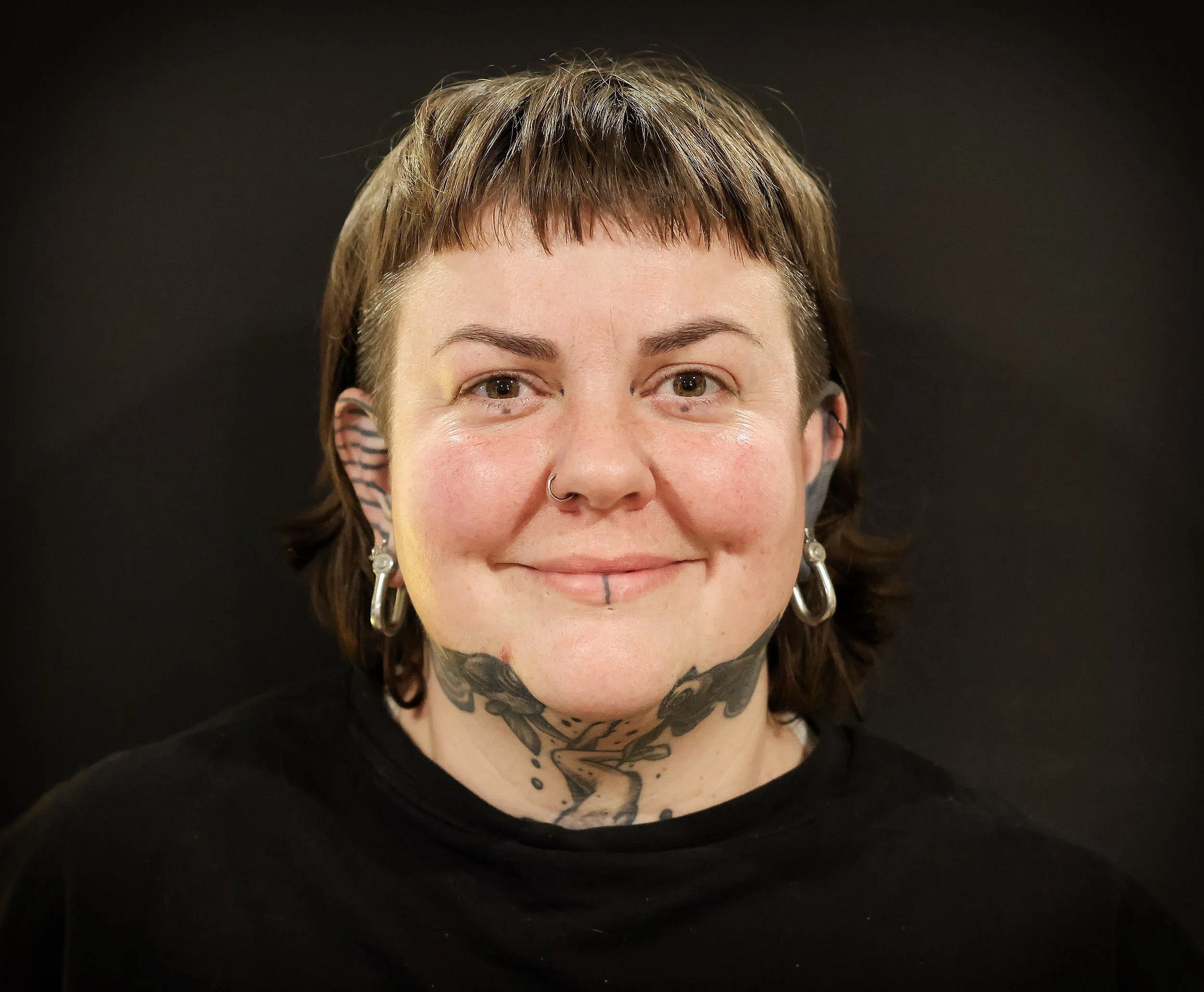 Close-up of a person with short brown hair, tattoos on neck and chest, and earrings, smiling against a dark background.