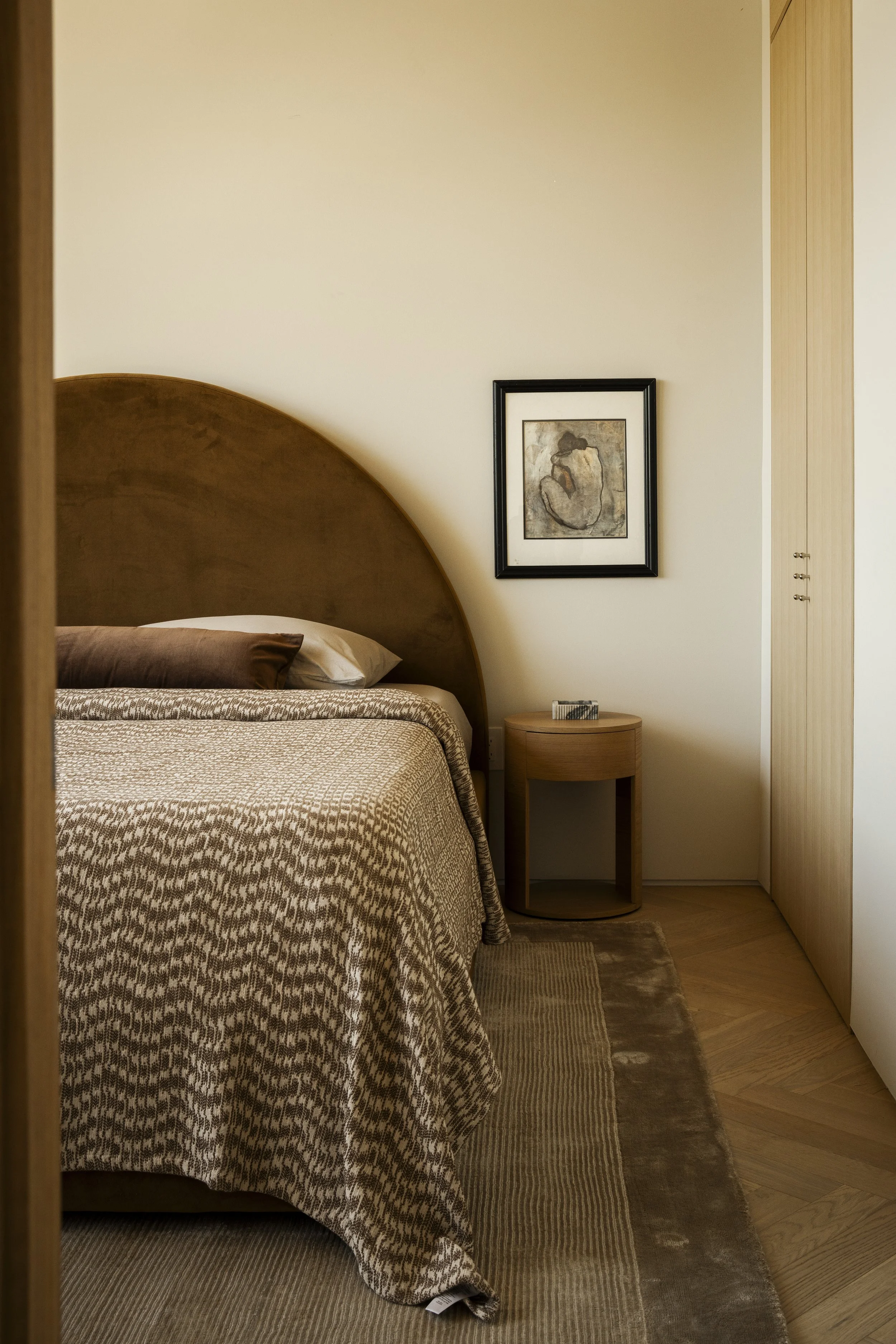 A bedroom corner with a bed, a framed abstract artwork on the wall, a small wooden nightstand with books, beige walls, wood flooring, and a rug.