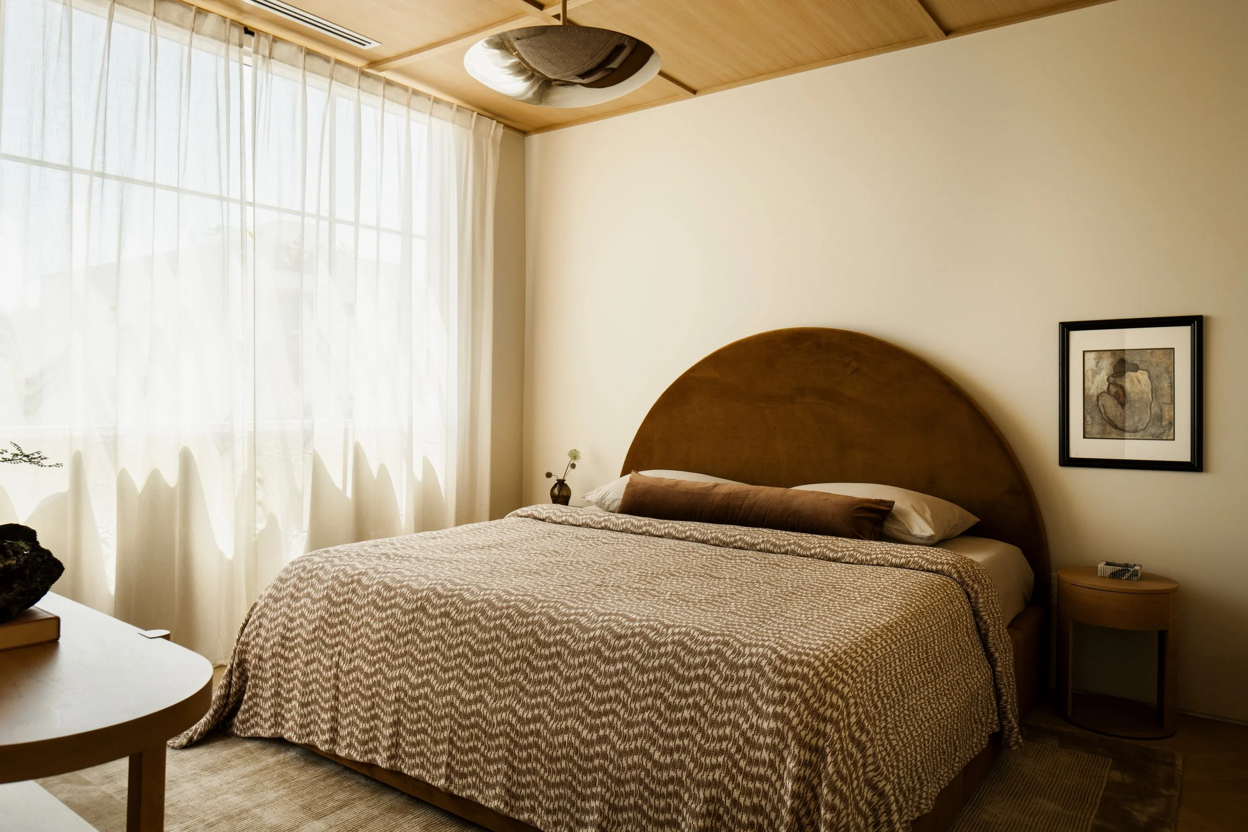 A bedroom with a large bed, wooden headboard, patterned beige bedspread, pillows, nightstand, framed artwork on the wall, and sheer curtains covering a window.