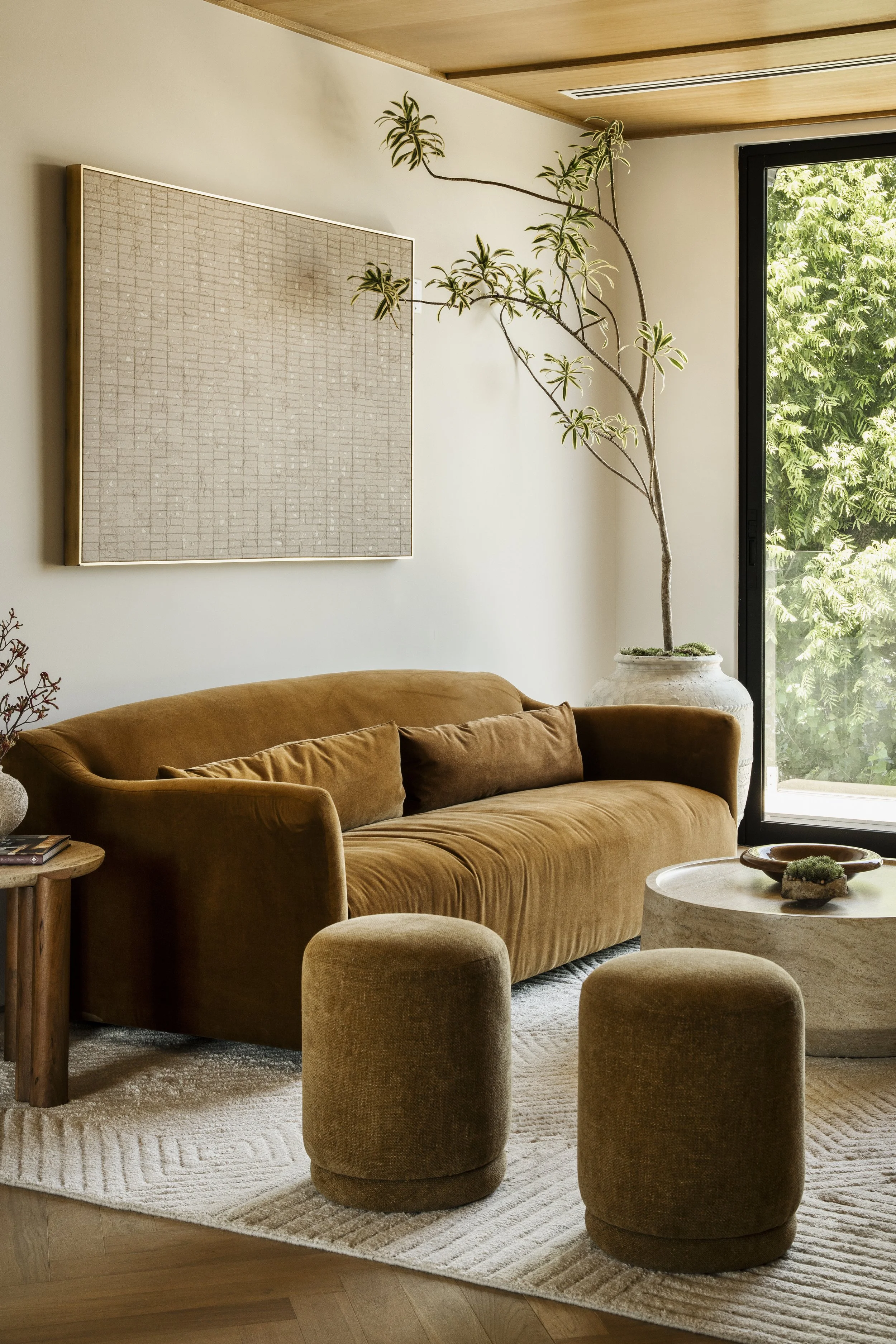 Living room with a tan velvet sofa, four matching ottomans, a round coffee table with decor, a large potted plant near a window, and a wall with abstract art.