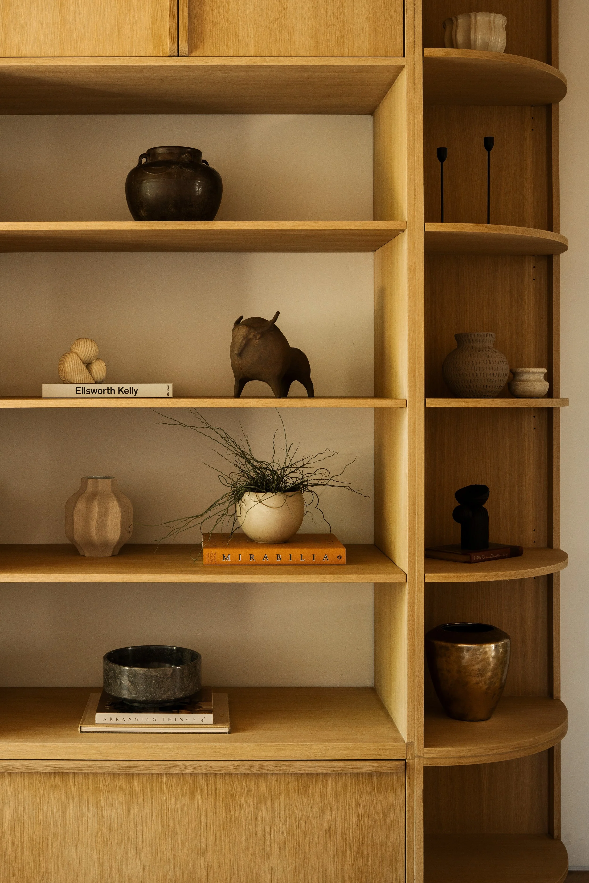 A wooden shelving unit with various decorative objects including pottery, a small sculpture of an animal, a plant, and books in a minimalist style.