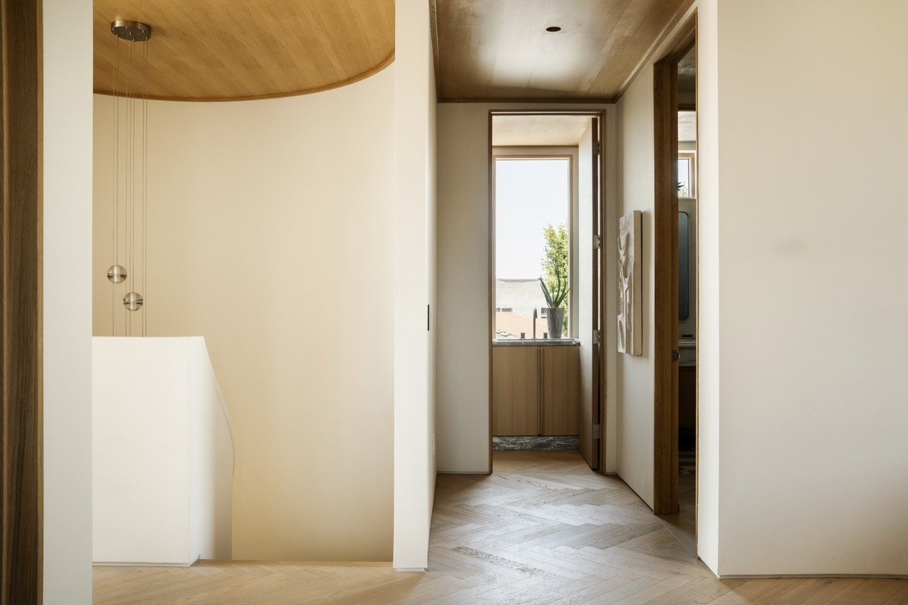 Interior view of a modern home hallway with wooden accents, white walls, a window with a potted plant on the sill, and hallway doors to the bedrooms or rooms.