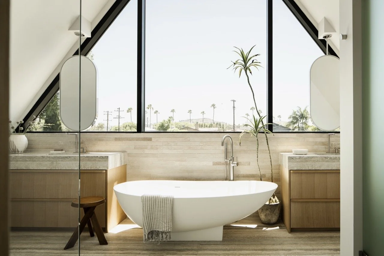 Modern bathroom with a freestanding bathtub, large window showing palm trees, two wooden vanities with sinks, plants, and mirrors.