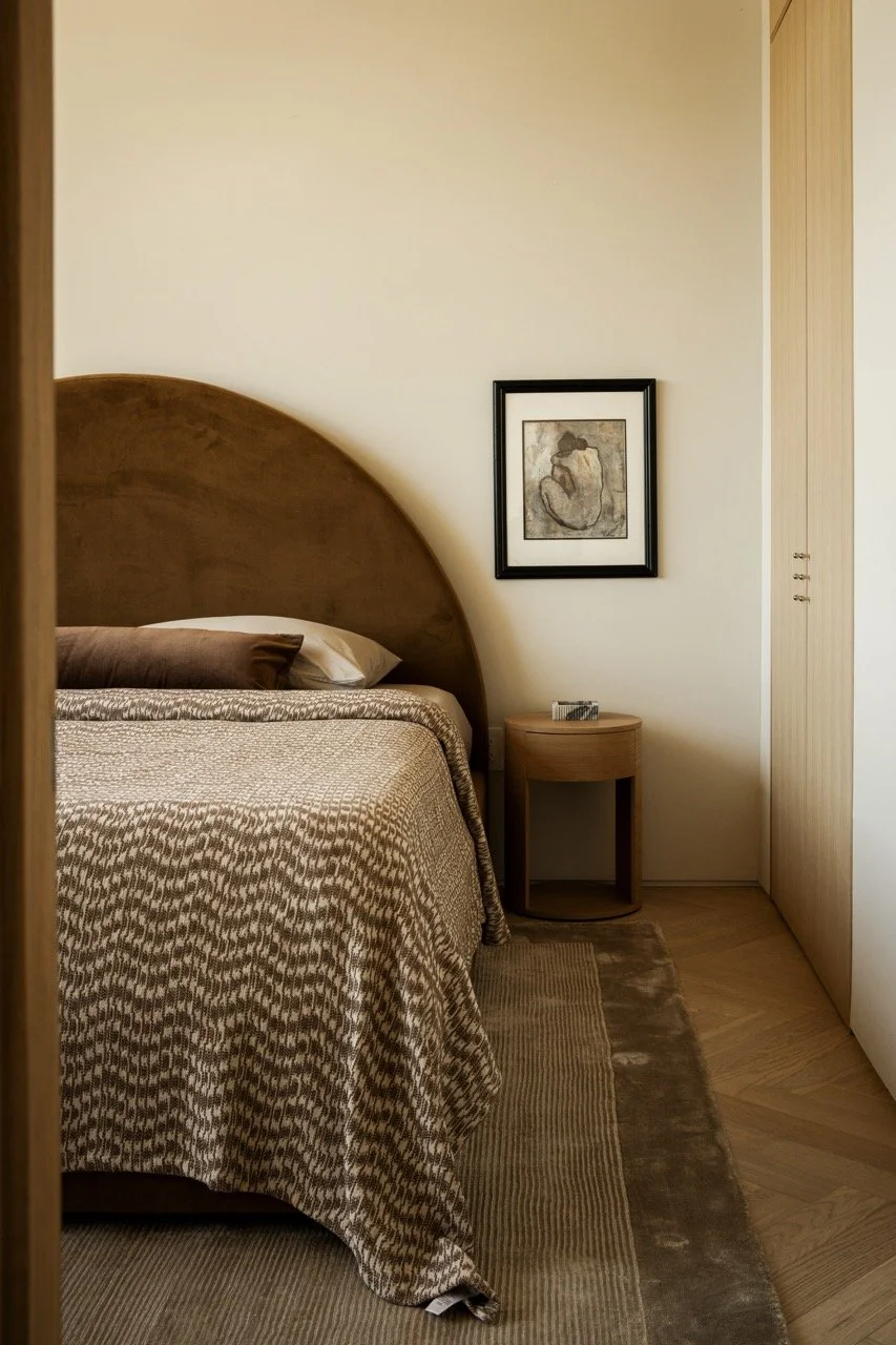 A bedroom with a wooden headboard, a bed with patterned bedding, a brown pillow, a rug, a small round bedside table, a framed artwork on the wall, and a closed closet door.