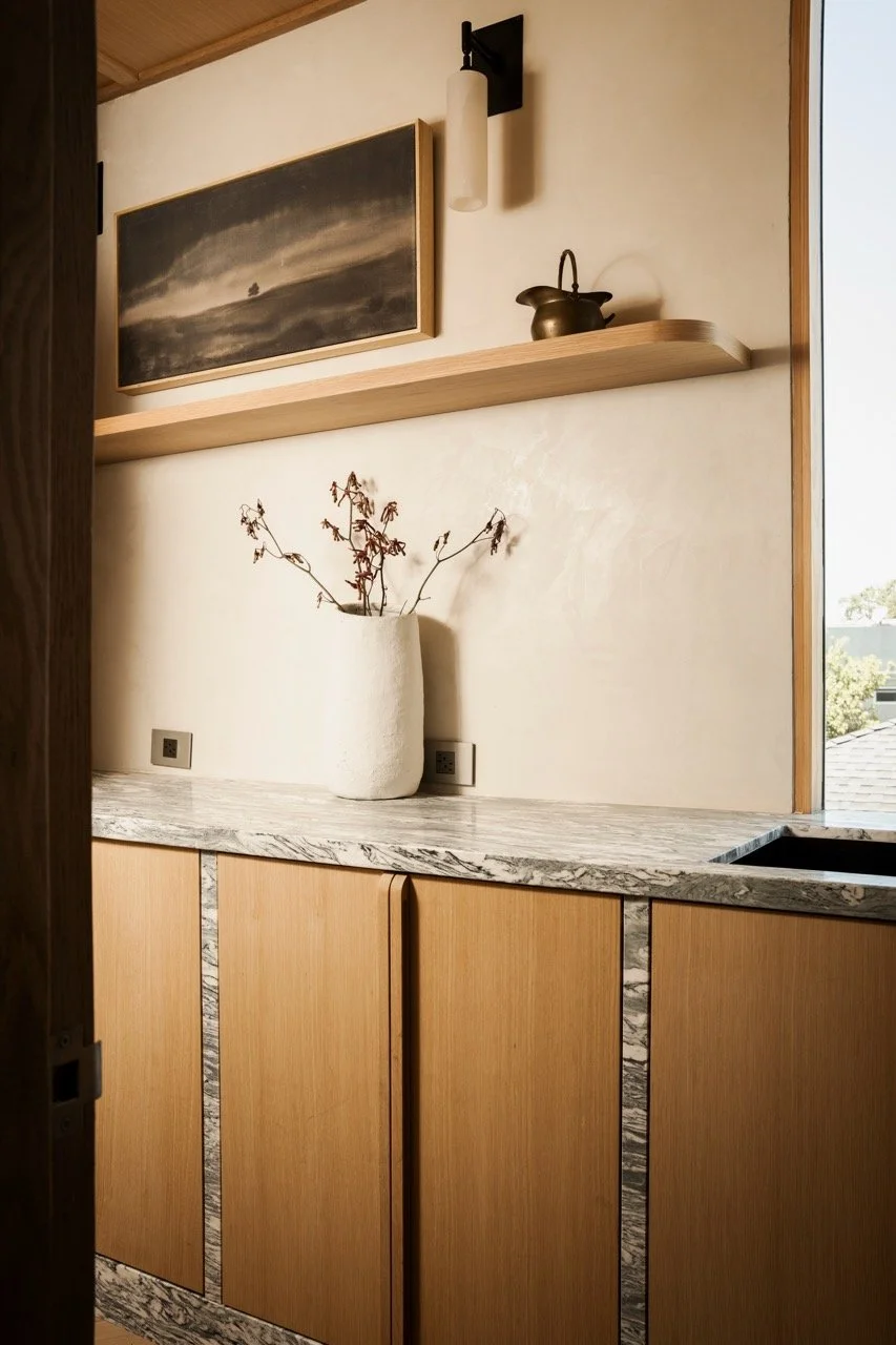 Interior of a modern kitchen with wooden cabinets, a marble countertop, a large white vase with dried branches, and a window with natural light.
