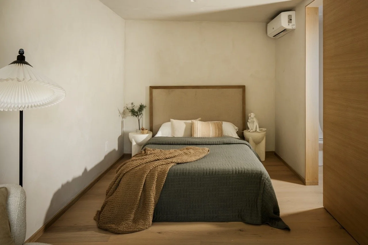 A cozy bedroom with a neatly made bed, beige headboard, and decorative pillows. Bed has a dark green coverlet and a brown throw blanket. Two white nightstands with decorative items and greenery sit on either side. An air conditioning unit is mounted 