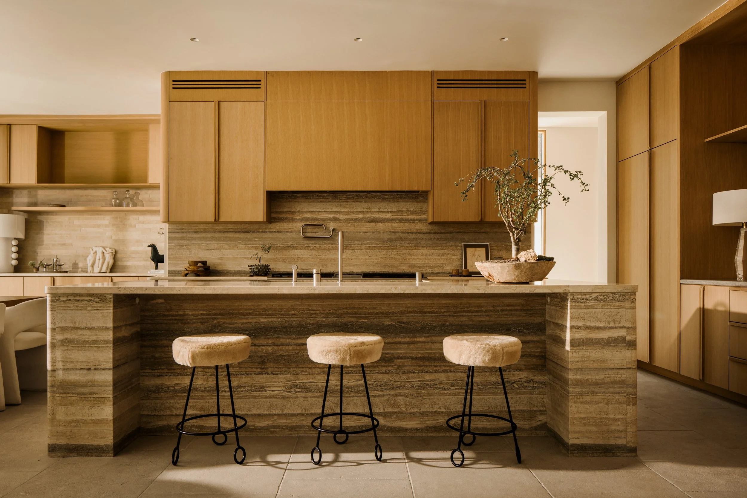 Modern kitchen with wood cabinetry, a marble island with three beige cushioned stools, small green plants on the counter, and a decorative bowl with rocks and a plant on the island.
