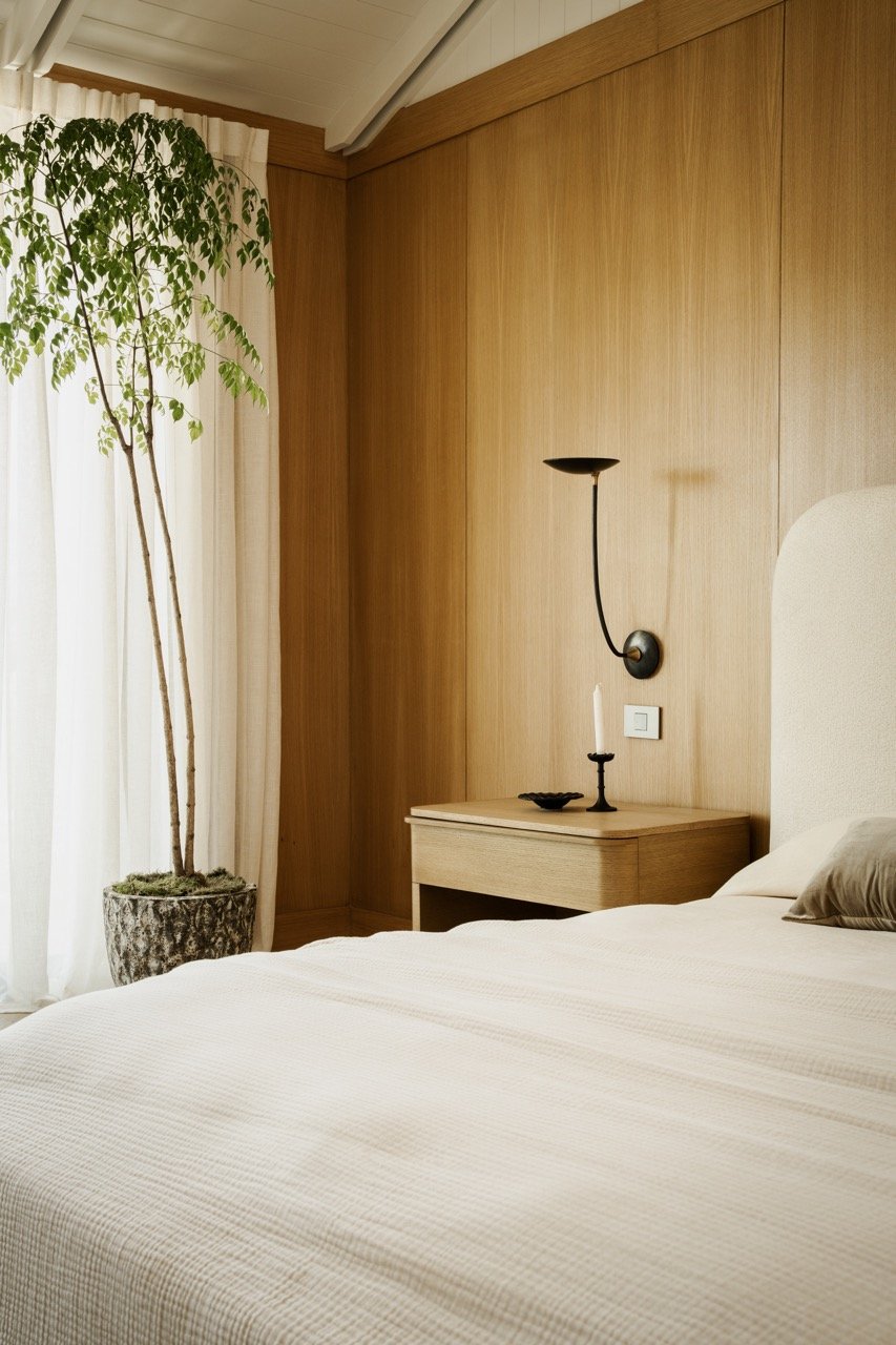 A bedroom corner with a bed, a wooden nightstand, a wall-mounted lamp, a candle holder with a candle, a large potted plant with green leaves, and cream-colored curtains.