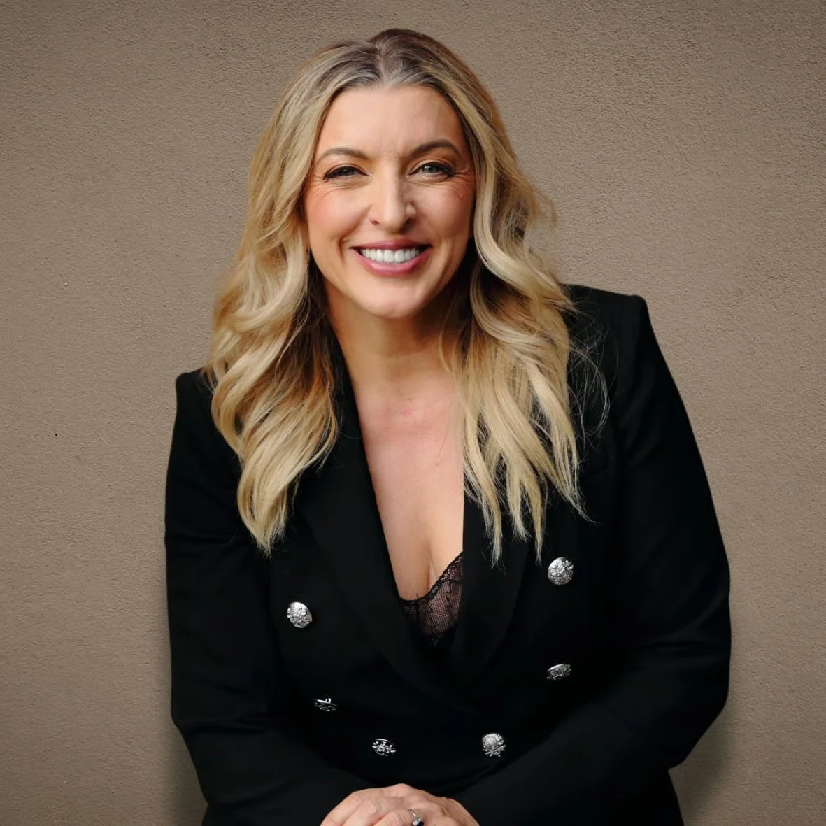 A smiling woman with blonde wavy hair wearing a black blazer with silver buttons against a neutral background.