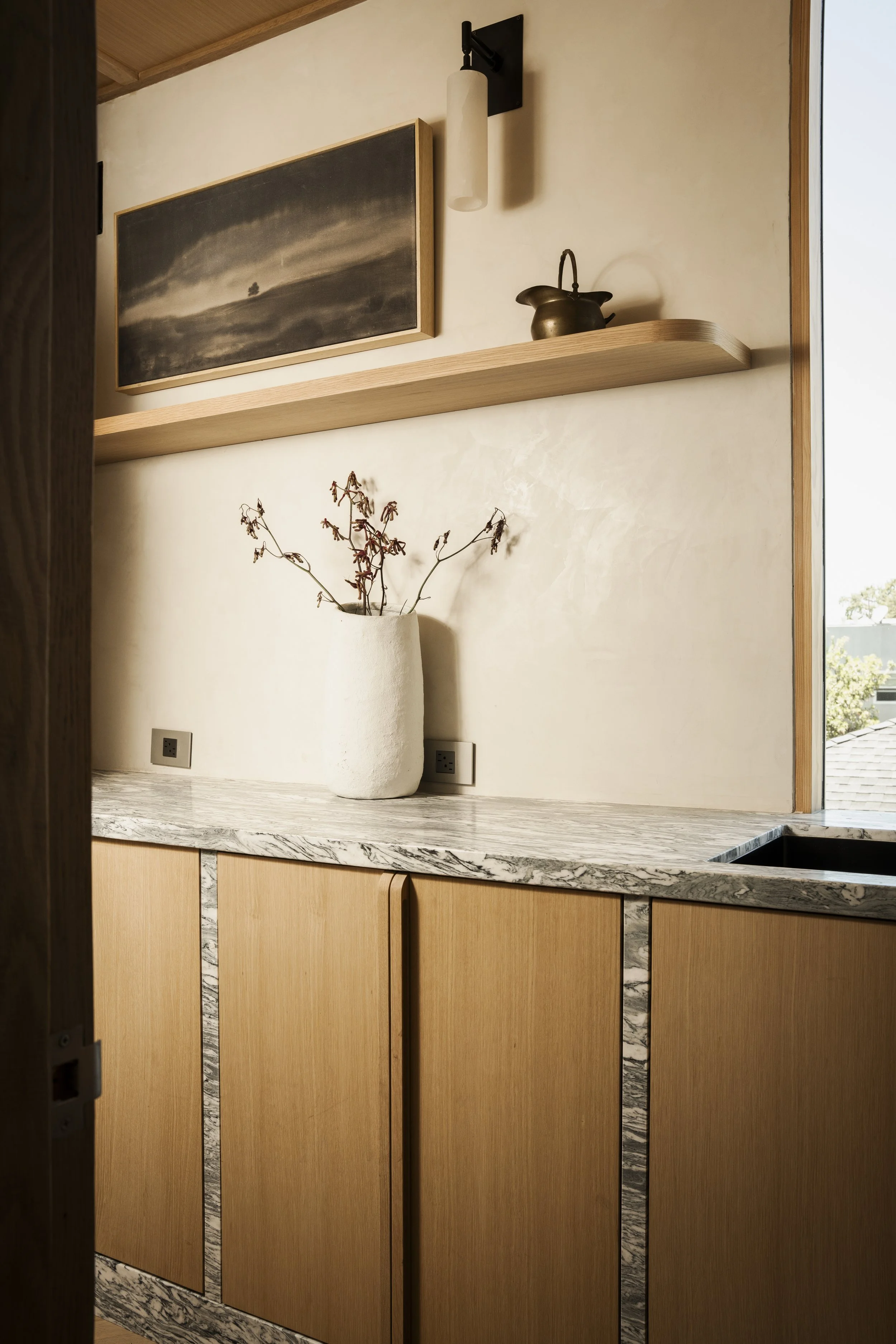 A kitchen with beige wooden cabinets, a marble countertop, a white vase with dried branches, a framed black and white landscape painting, a wooden shelf with a vintage teapot, and electrical outlets.