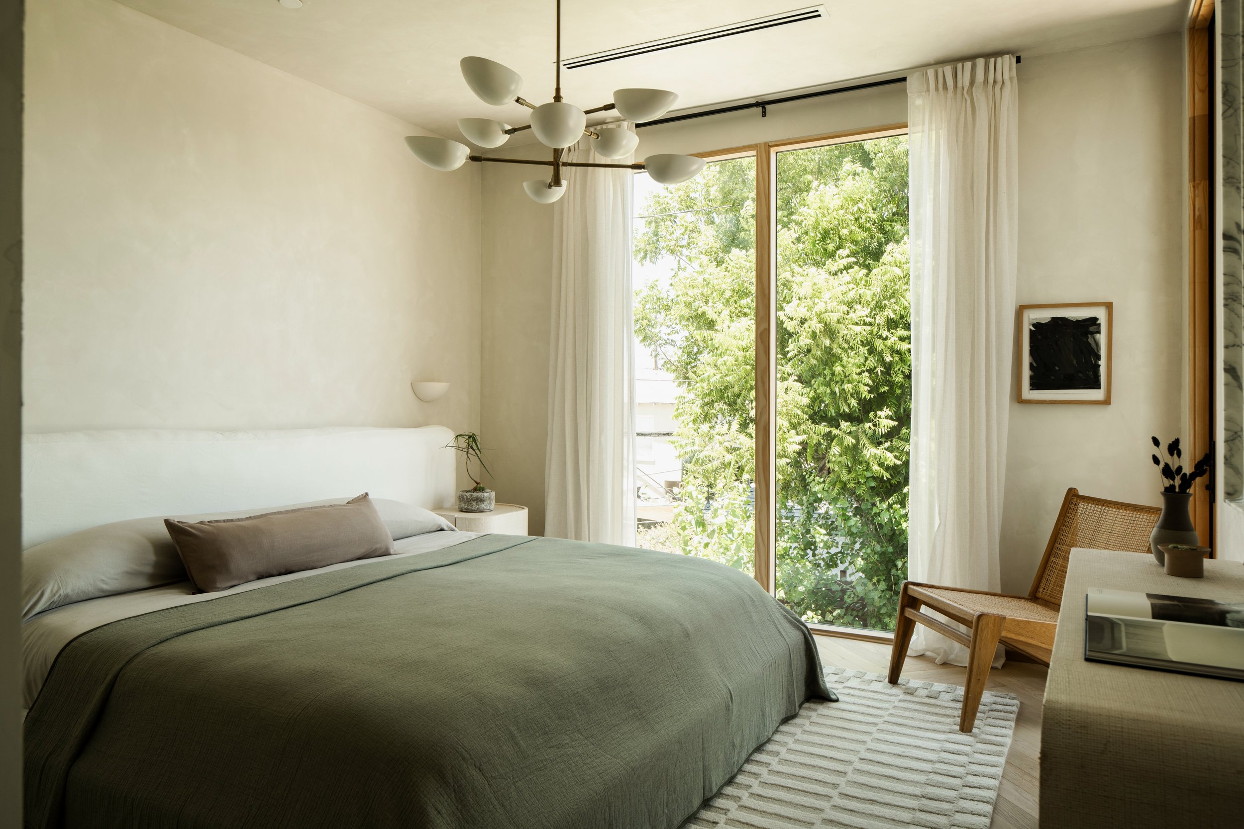 A cozy bedroom with a large bed covered in a green blanket and beige pillows. A nightstand with a potted plant is next to the bed. Large windows with white curtains reveal lush green trees outside. A modern chandelier hangs from the ceiling, and ther