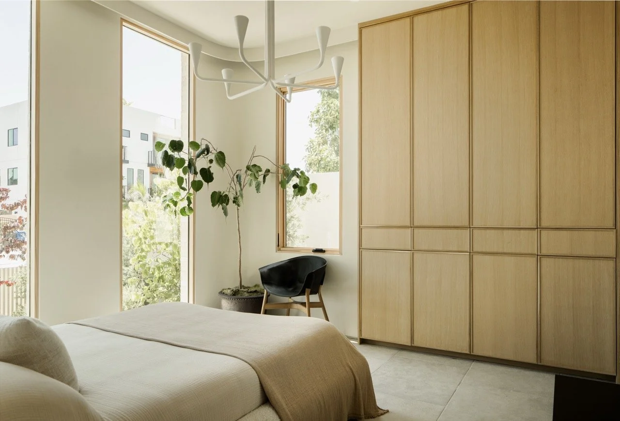 A bedroom with large windows, a bed with beige bedding, a black chair, a potted houseplant, a modern white chandelier, and a large wooden wardrobe.