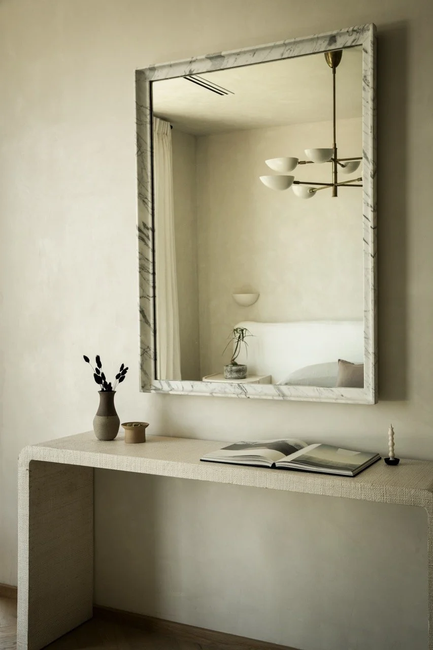 Decorative mirror hanging above a minimalist cream-colored console table with a small flower arrangement, an open book, and a candle.