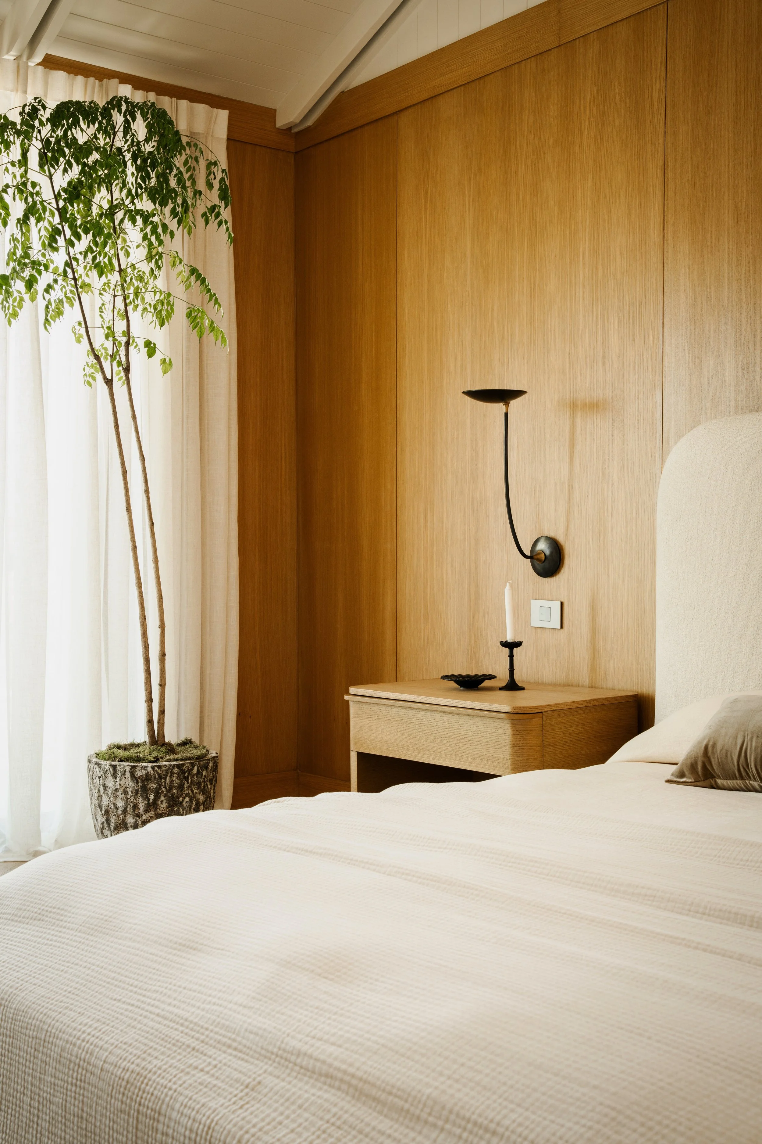 A bedroom featuring a bed with a white textured cover, a bedside table with a lamp and candle holder, a large potted plant near curtain-covered window, and a wooden wall background.
