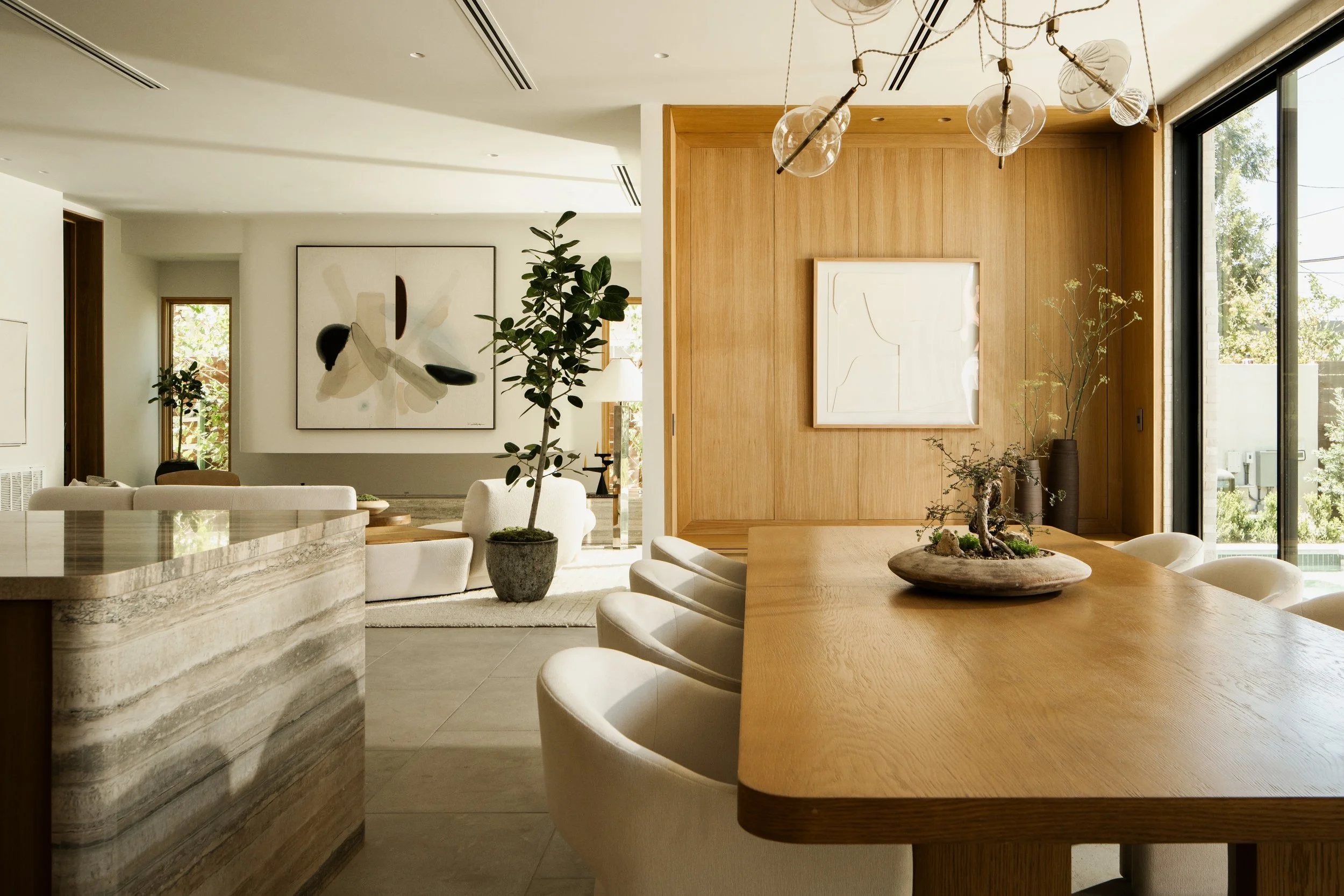 Modern living room with wooden accents, a large window, indoor plants, artwork on the walls, and a kitchen island in the foreground.