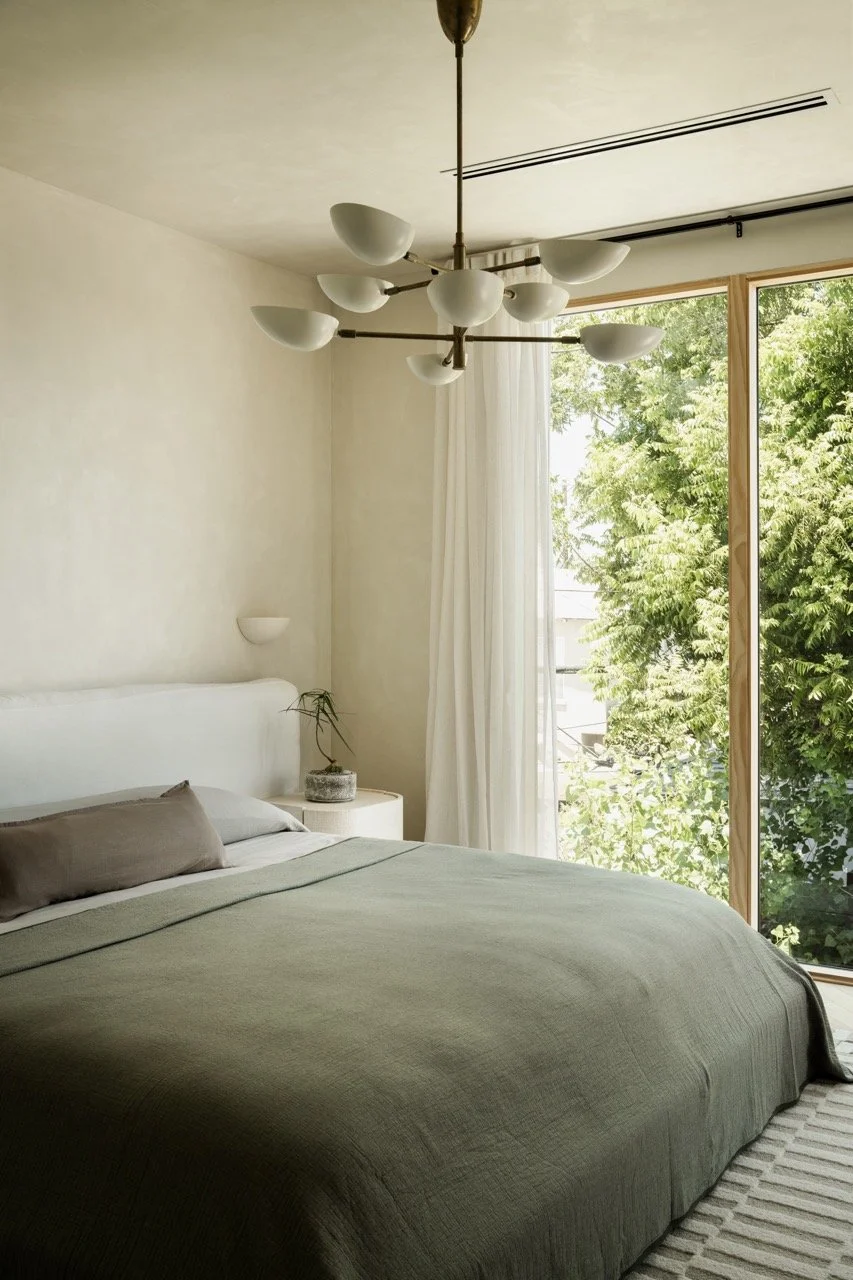 A minimalist bedroom with a bed covered in an olive green bedspread, a white headboard, a taupe pillow, a small white bedside table with a potted plant, a modern chandelier with white bowls, white curtains, and a large window showing green trees outs