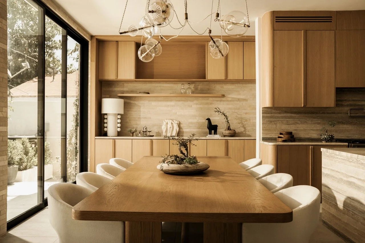 Modern dining room with a large wooden table surrounded by white chairs, decorated with a small bonsai tree. The room features light wood cabinetry, a sliding glass door, and contemporary light fixtures hanging from the ceiling.