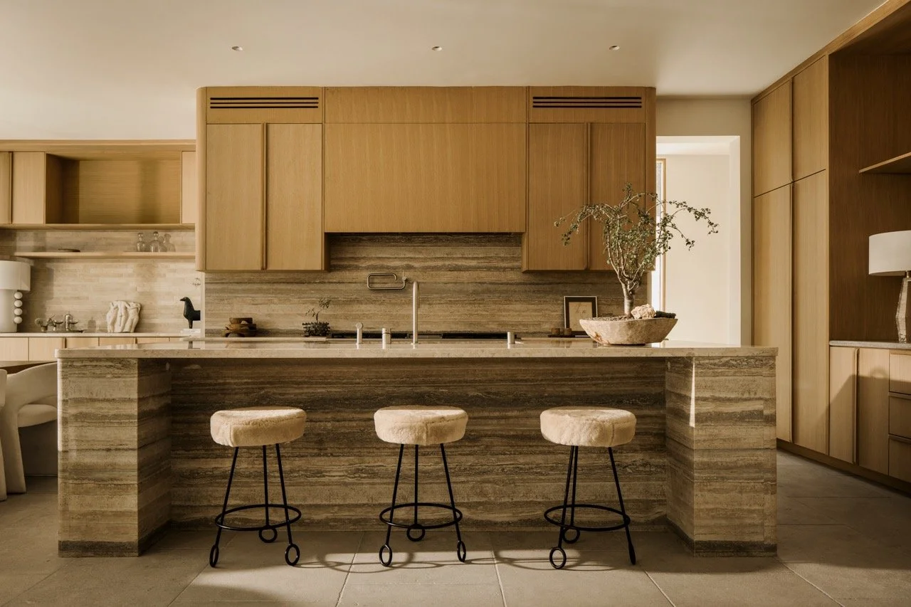 Modern kitchen with a marble island, three beige bar stools, wooden cabinets, and a potted plant.