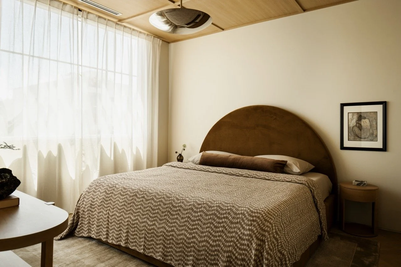 A bedroom with a large bed, brown headboard, beige patterned bedspread, and pillows. There is a small nightstand with a vase and flower, a framed picture on the wall, and sheer curtains covering a window. The ceiling has a wooden finish with a circul