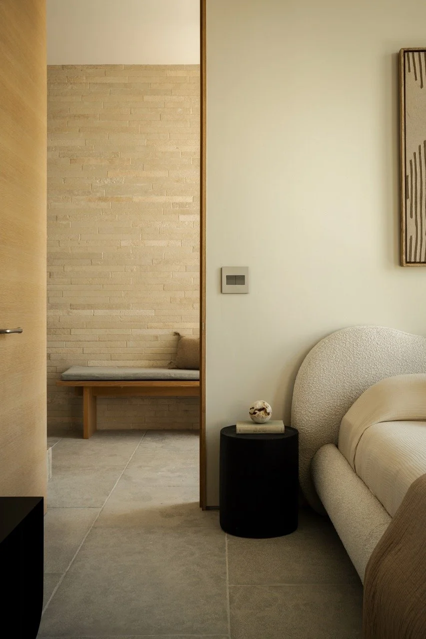 Interior view of a modern bedroom with a beige bed, black side table with decorative object, minimal wall art, and a corner seating area with a bench and cushion.