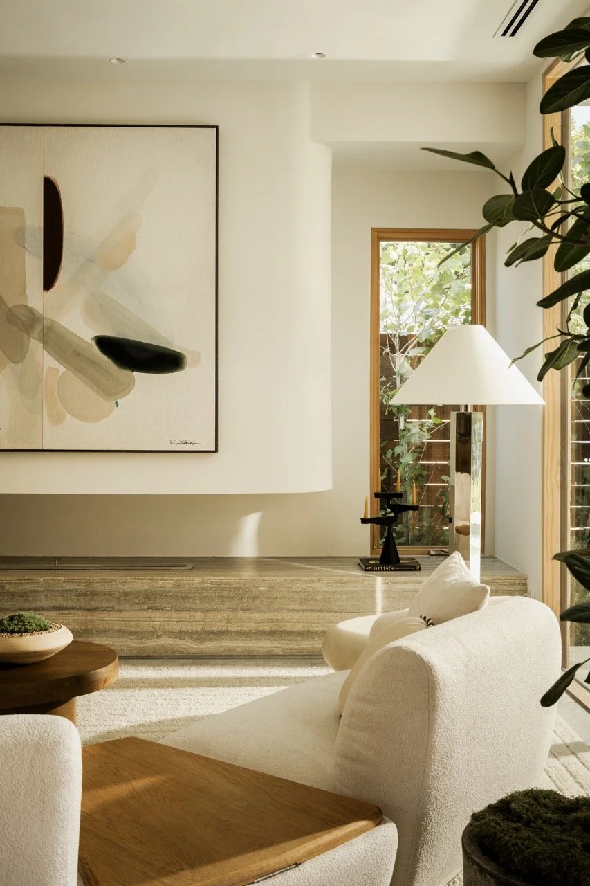 Bright living room with white sofa, wooden side table, large abstract painting, floor lamp, and large windows with greenery outside.