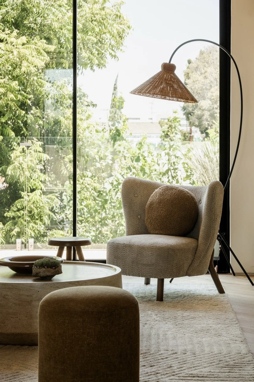 A cozy living room corner with a beige armchair, a round cushion, and a wood coffee table in front. A large floor lamp with a wicker shade stands beside the chair. Floor-to-ceiling windows reveal greenery outside, filling the room with natural light.