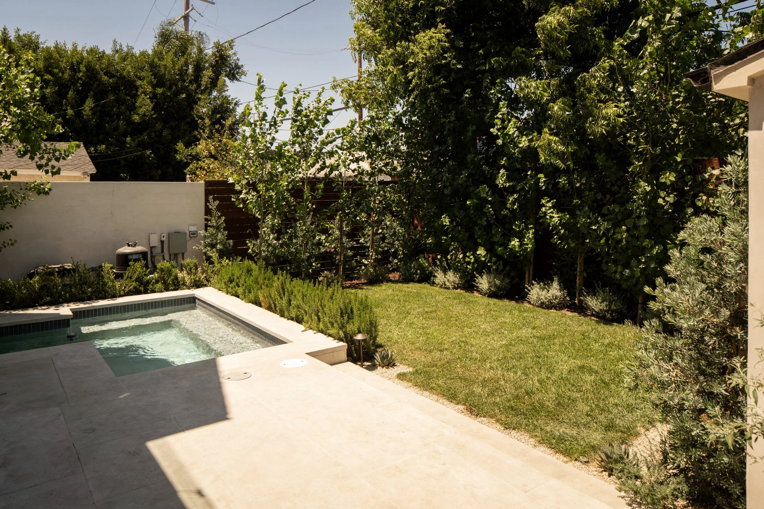 Backyard with swimming pool, green lawn, shrubs, and tall trees. A white fence and utility boxes are visible, with power lines overhead, under a clear sky.