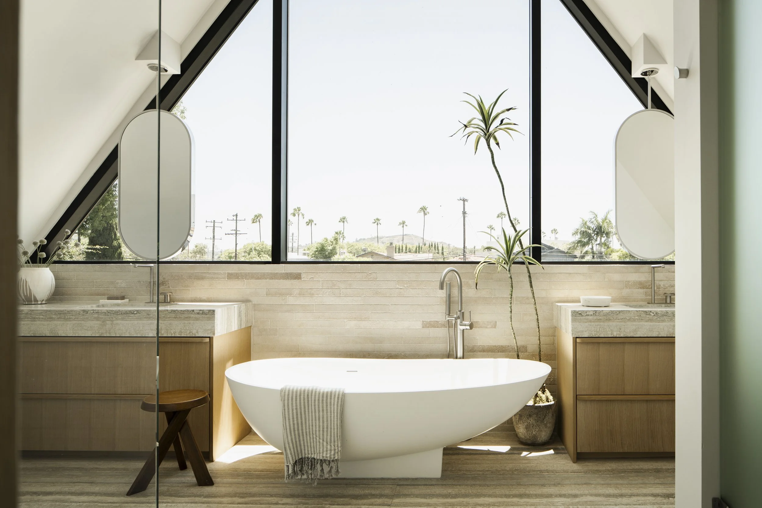 Modern bathroom with large window, bathtub, wooden vanity, potted plants, and minimal decor.