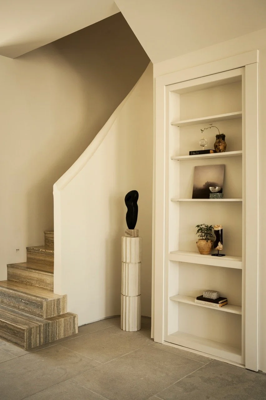 Interior corner of a home with a staircase, a pedestal with a sculpture, and a built-in white bookshelf with decorative objects and books.