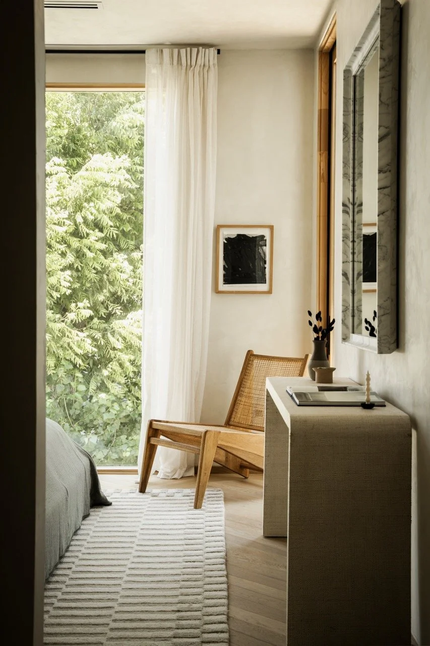 A corner of a bedroom with a large window covered by white curtains, a wooden chair, a light-colored rug, and a white dresser with decor items.