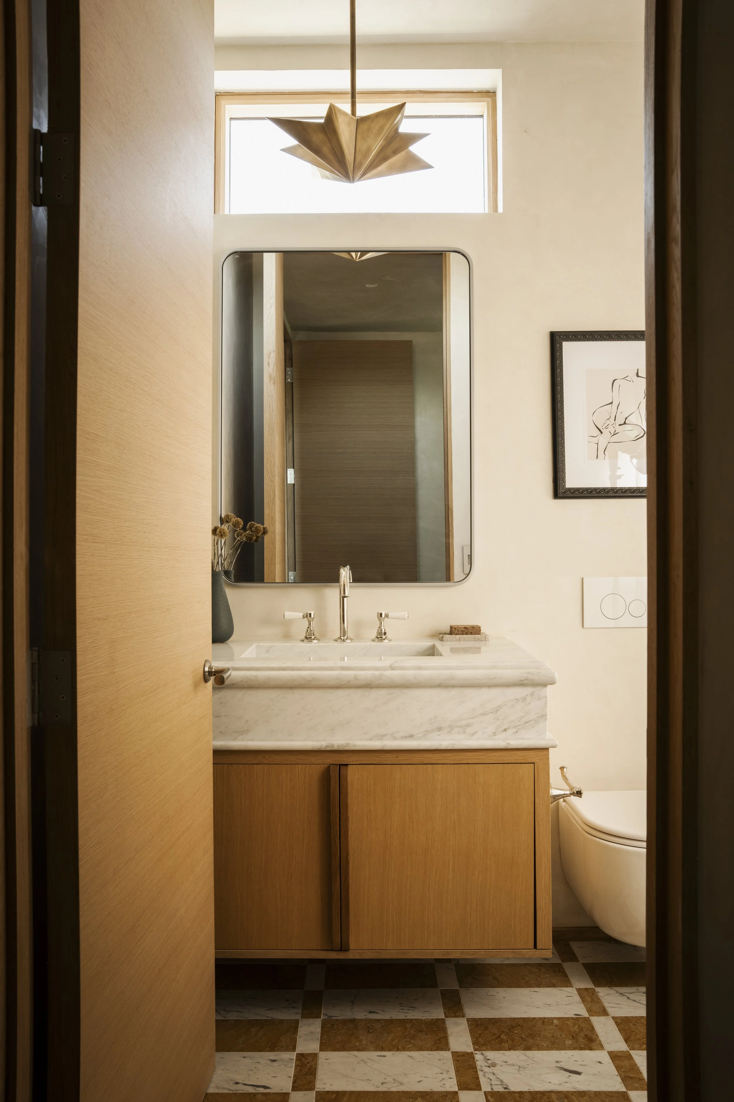 A bathroom with a wooden vanity, a marble countertop, a wall mirror, a vase with dried flowers, framed artwork, a window, and a modern toilet with a rounded design.