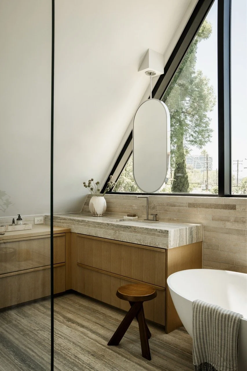 Modern bathroom with a large sloped window, wooden cabinetry, a freestanding bathtub, a vase with dried flowers, and a tall mirror.