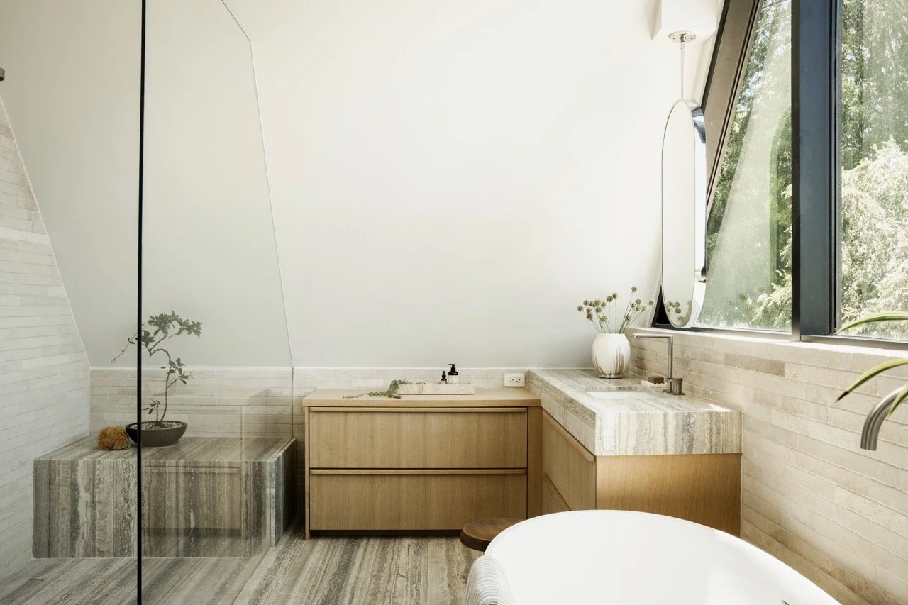 Modern bathroom with a wooden vanity, marble countertop sink, large window with greenery outside, round mirror, potted plant, decorative vase, and minimalist decor.