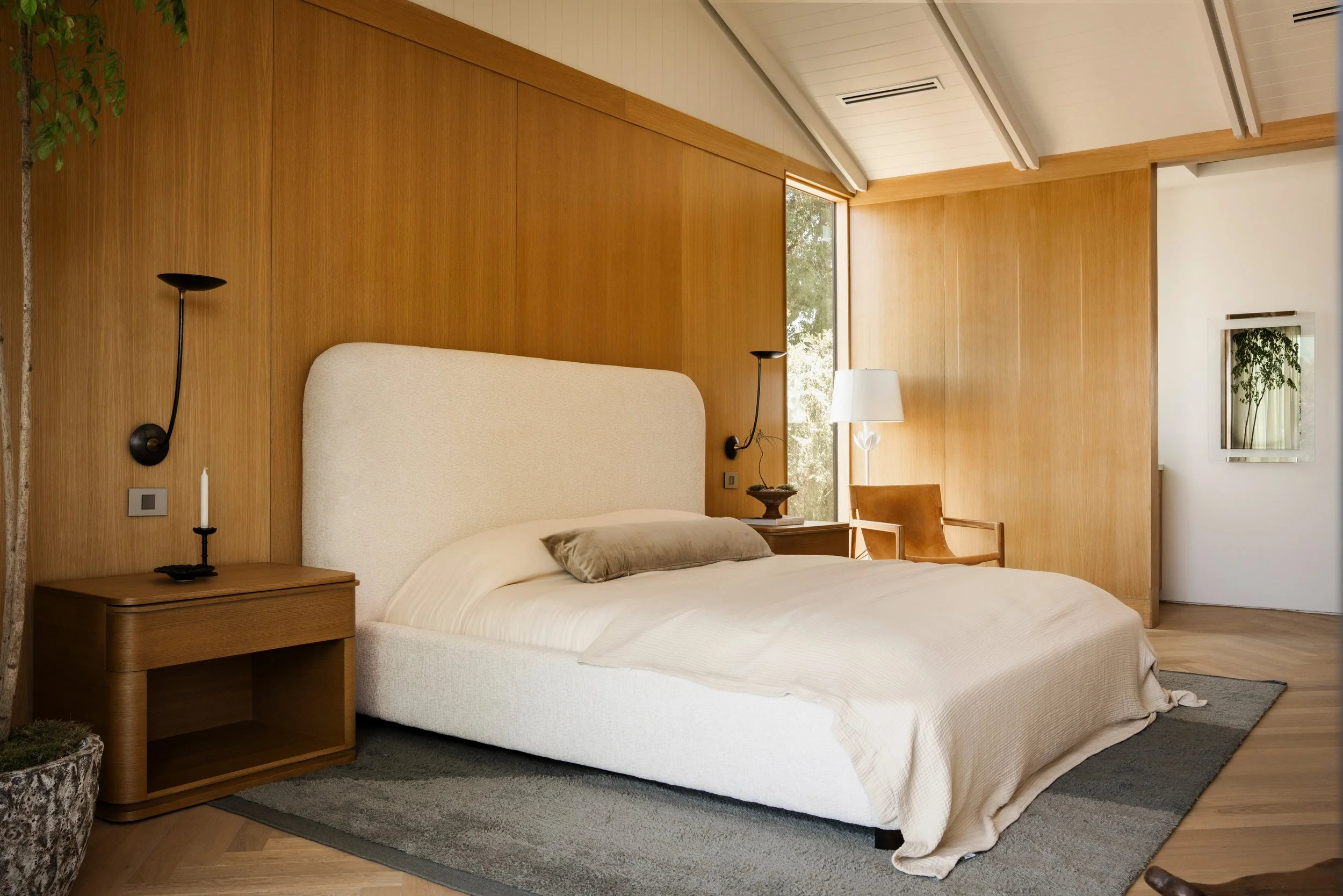 A bedroom with a large bed featuring a beige upholstered headboard, a wooden nightstand with a black lamp, a white chair, a table lamp, a window with natural light, wooden wall paneling, and a framed picture with plants on the white wall.