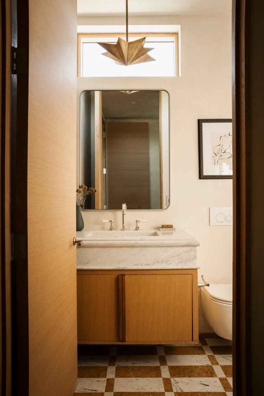 View of a modern bathroom through an open wooden door, featuring a marble countertop sink, a large mirror, a framed abstract line drawing on the wall, and a contemporary wall-mounted toilet, with a geometric pendant light and a small window above.