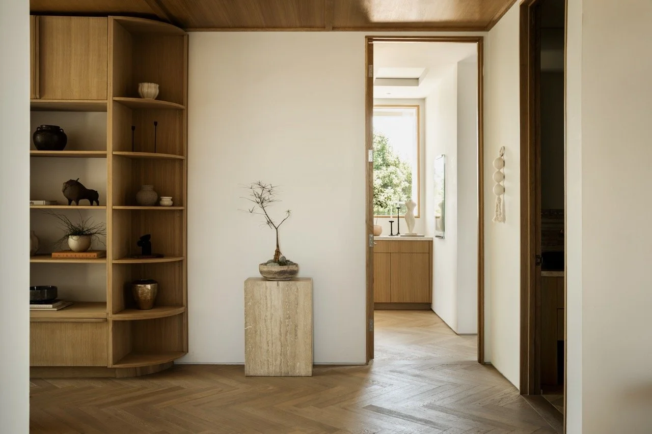 Minimalist interior with a wooden bookshelf on the left filled with decorative items, a small potted bonsai plant on a beige stone pedestal in the center, and a doorway leading to a Sunlit room with a window, a white vase, and decorative candles.