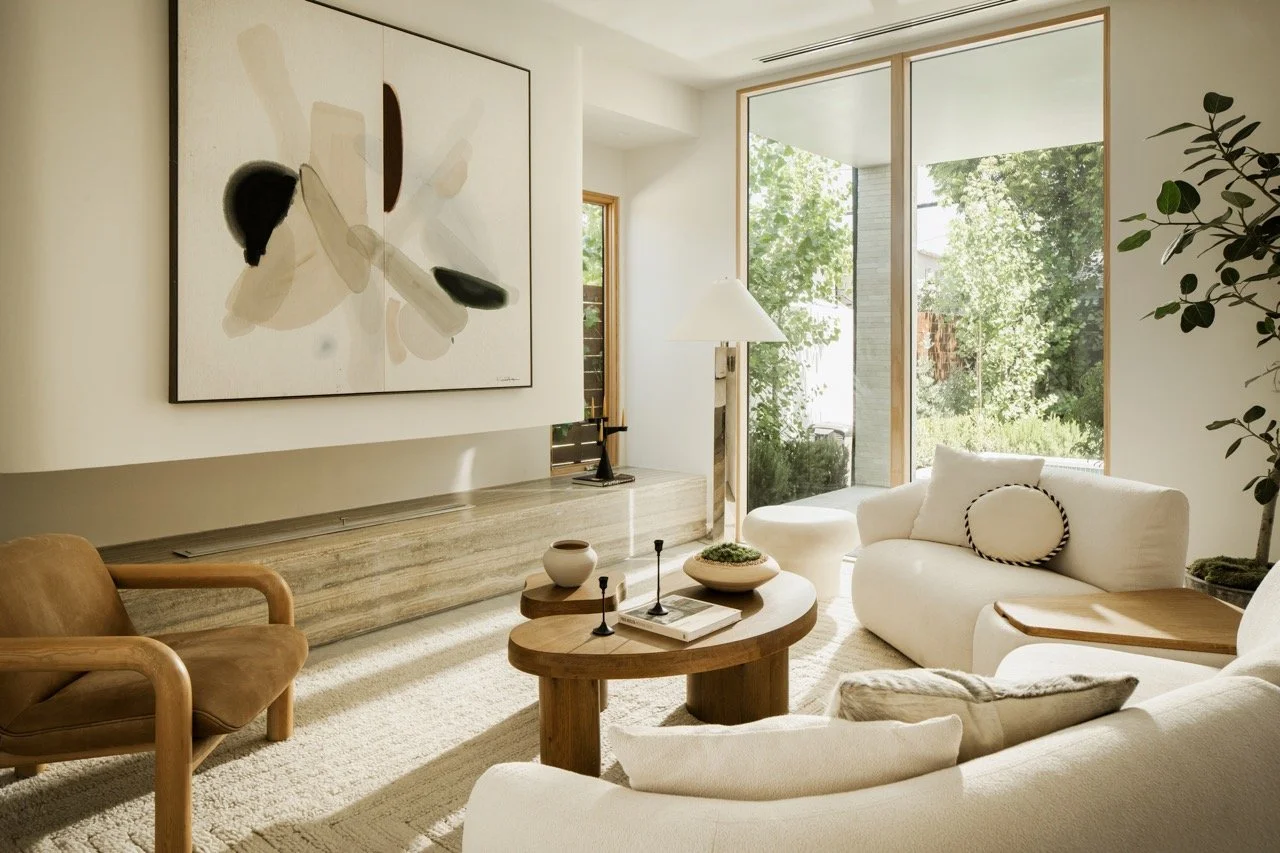 Bright living room with large windows, abstract wall art, beige sofas, a wooden coffee table, and green plants.
