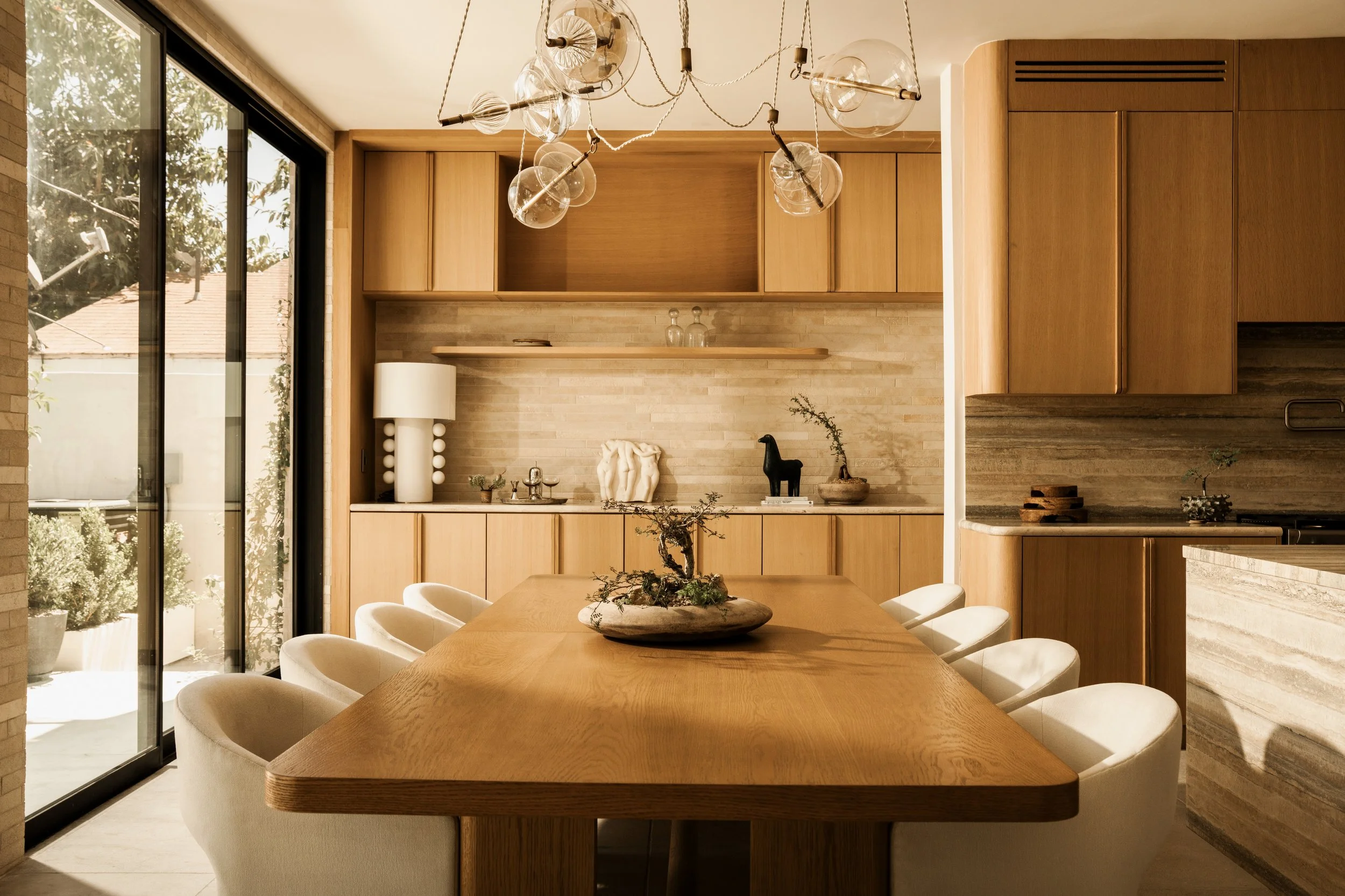 Modern kitchen and dining area with wooden cabinets, a large wooden table, white chairs, and unique pendant lighting.