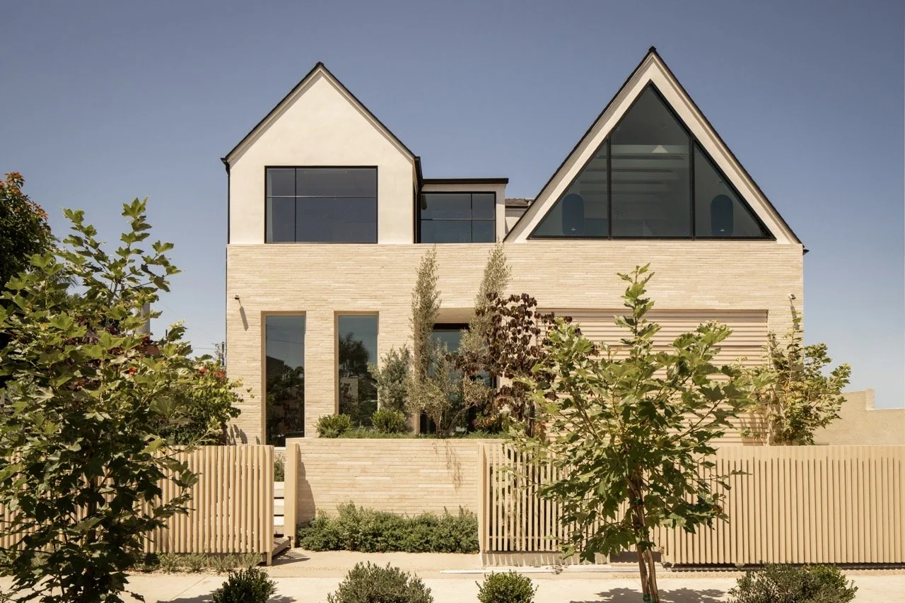 Modern house with sloped and triangular windows, surrounded by greenery and wooden fencing.