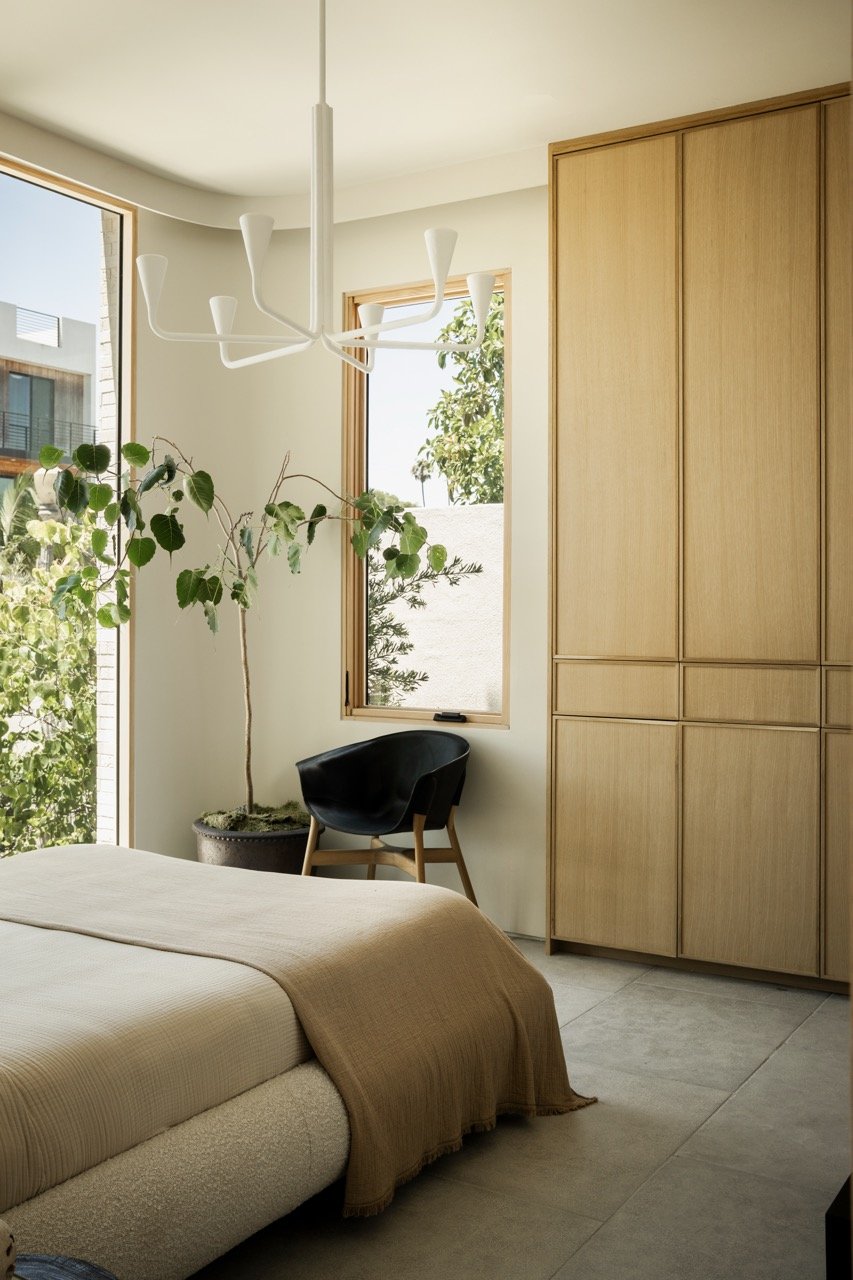 A bedroom with a beige bed, a large window with wooden trim letting in natural light, a tall potted plant, a black chair, and a wooden closet.