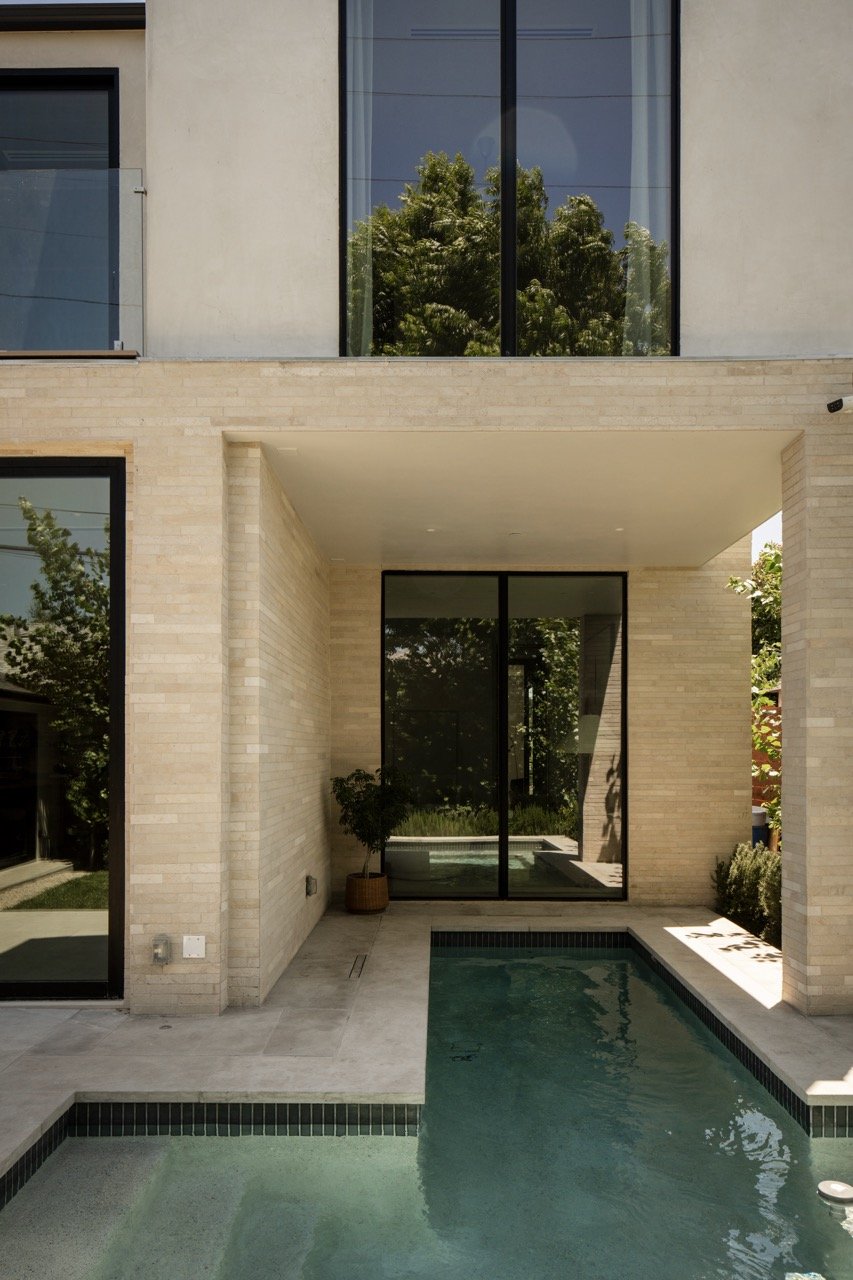 Modern house with large glass windows and a small swimming pool outside, surrounded by beige brick walls and greenery.