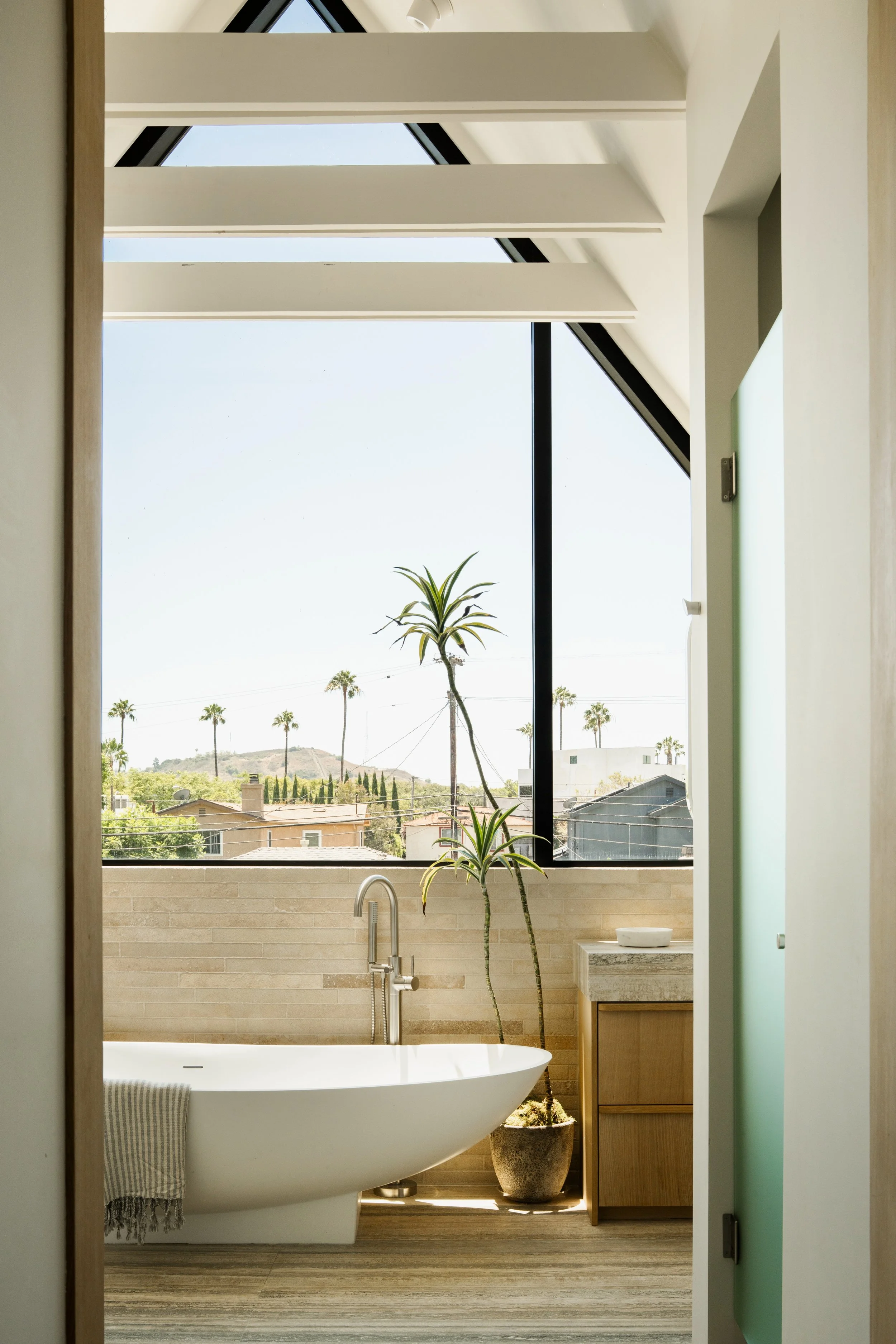 A modern bathroom with a freestanding bathtub, a window with a view of palm trees, a potted plant, and a small sink area.