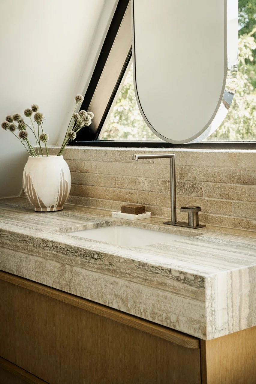 Close-up of a modern bathroom sink with a beige marble countertop, a chrome faucet, a small soap dish with soap, a decorative vase with dried flowers, and a round mirror above the sink, with natural light and greenery visible through a window.